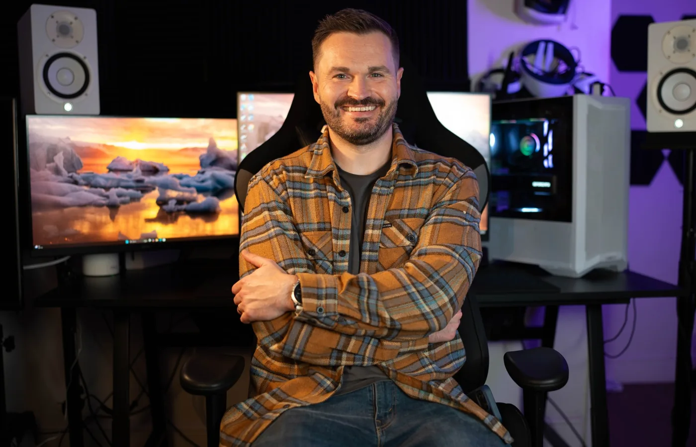 Sam Buckingham smiling with arms folded at a desk with monitors, studio speakers, a PC and VR headsets, lit in purple